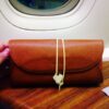 A brown leather pouch is placed on a wooden surface in front of an airplane window. The pouch is secured with a yellow string and a small white tag. The background shows a blurred view outside the airplane window.