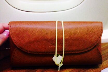 A brown leather pouch is placed on a wooden surface in front of an airplane window. The pouch is secured with a yellow string and a small white tag. The background shows a blurred view outside the airplane window.