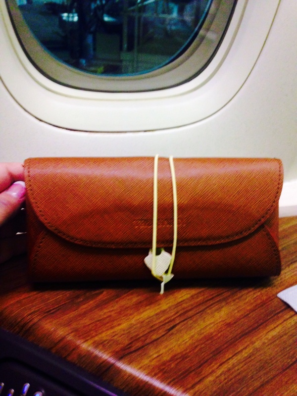 A brown leather pouch is placed on a wooden surface in front of an airplane window. The pouch is secured with a yellow string and a small white tag. The background shows a blurred view outside the airplane window.