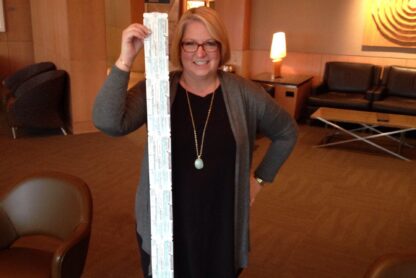 A woman is standing in a room holding up a long strip of what appears to be medical or prescription labels. She is smiling and wearing glasses, a black dress, and a gray cardigan. The room has modern decor with brown chairs, wooden walls, and a lit lamp in the background.