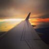 The image shows the wing of an airplane in flight, with a view of a sunset or sunrise in the background. The sky is filled with a mix of warm colors, including orange, yellow, and hints of blue, reflecting off a body of water below. The horizon is visible, with some landmasses and clouds adding to the scenic view.