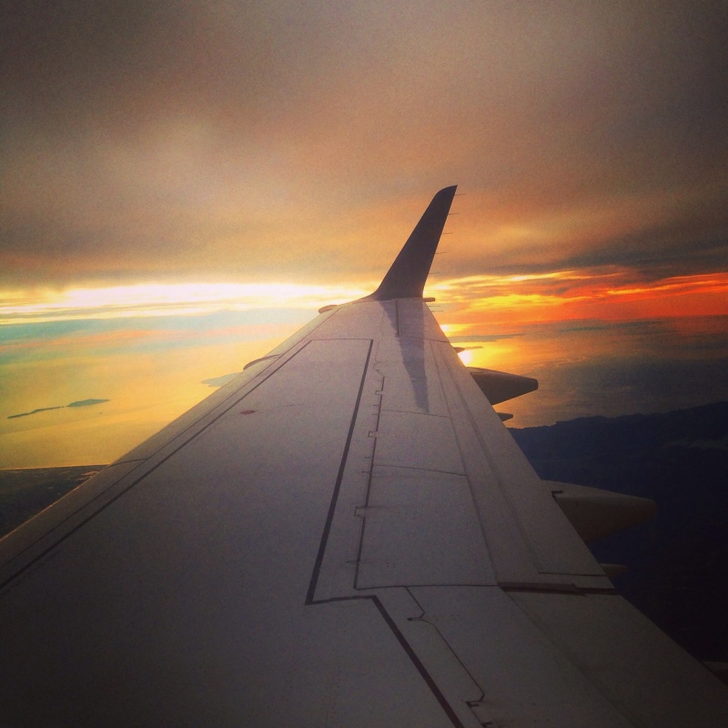 The image shows the wing of an airplane in flight, with a view of a sunset or sunrise in the background. The sky is filled with a mix of warm colors, including orange, yellow, and hints of blue, reflecting off a body of water below. The horizon is visible, with some landmasses and clouds adding to the scenic view.