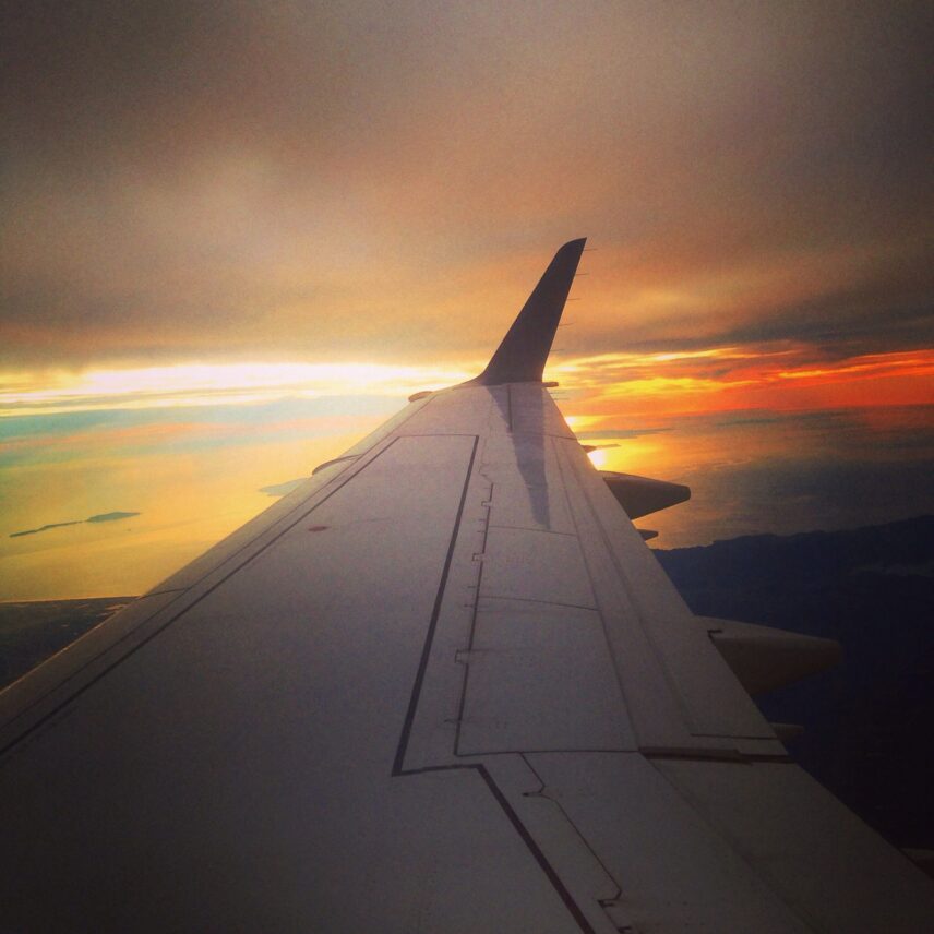 The image shows the wing of an airplane in flight, with a view of a sunset or sunrise in the background. The sky is filled with a mix of warm colors, including orange, yellow, and hints of blue, reflecting off a body of water below. The horizon is visible, with some landmasses and clouds adding to the scenic view.