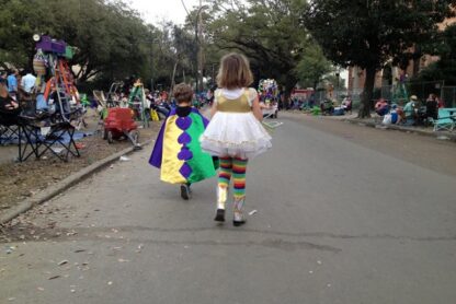 Two children are walking down a street during a parade or festival. One child is wearing a colorful cape with purple, green, and yellow sections, while the other child is dressed in a white dress with gold accents and rainbow-striped leggings. The street is lined with people sitting on chairs and ladders, and there are trees and decorations in the background.