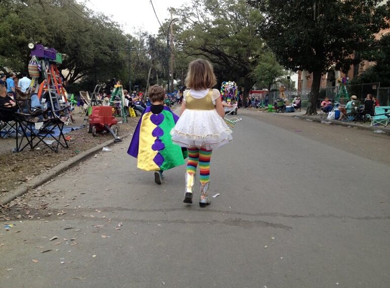 Two children are walking down a street during a parade or festival. One child is wearing a colorful cape with purple, green, and yellow sections, while the other child is dressed in a white dress with gold accents and rainbow-striped leggings. The street is lined with people sitting on chairs and ladders, and there are trees and decorations in the background.