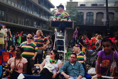 A group of people is gathered outdoors, likely at a parade or festival. Many are wearing colorful, striped shirts and beaded necklaces, suggesting a Mardi Gras celebration. Some are seated in folding chairs, while others are standing. A person is elevated on a ladder seat, providing a better view of the event. The background shows multi-story buildings and a partly cloudy sky. The atmosphere appears festive and lively.