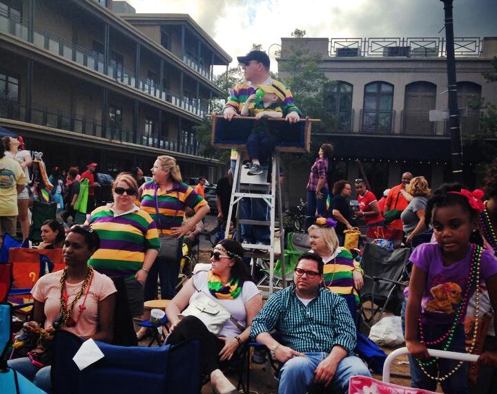 A group of people is gathered outdoors, likely at a parade or festival. Many are wearing colorful, striped shirts and beaded necklaces, suggesting a Mardi Gras celebration. Some are seated in folding chairs, while others are standing. A person is elevated on a ladder seat, providing a better view of the event. The background shows multi-story buildings and a partly cloudy sky. The atmosphere appears festive and lively.