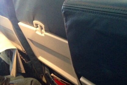 The image shows the interior of an airplane cabin. The focus is on the back of two seats, with passengers seated in them. The seats are upholstered in dark blue leather. The seatback pockets contain magazines and safety information cards. One passenger's hand is visible resting on their lap, and another passenger is seen wearing a cap, looking down. The overall scene suggests a typical in-flight setting.
