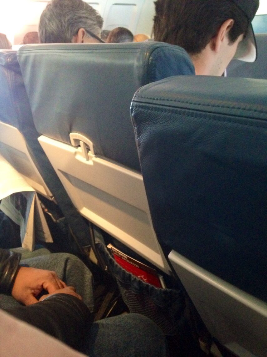 The image shows the interior of an airplane cabin. The focus is on the back of two seats, with passengers seated in them. The seats are upholstered in dark blue leather. The seatback pockets contain magazines and safety information cards. One passenger's hand is visible resting on their lap, and another passenger is seen wearing a cap, looking down. The overall scene suggests a typical in-flight setting.