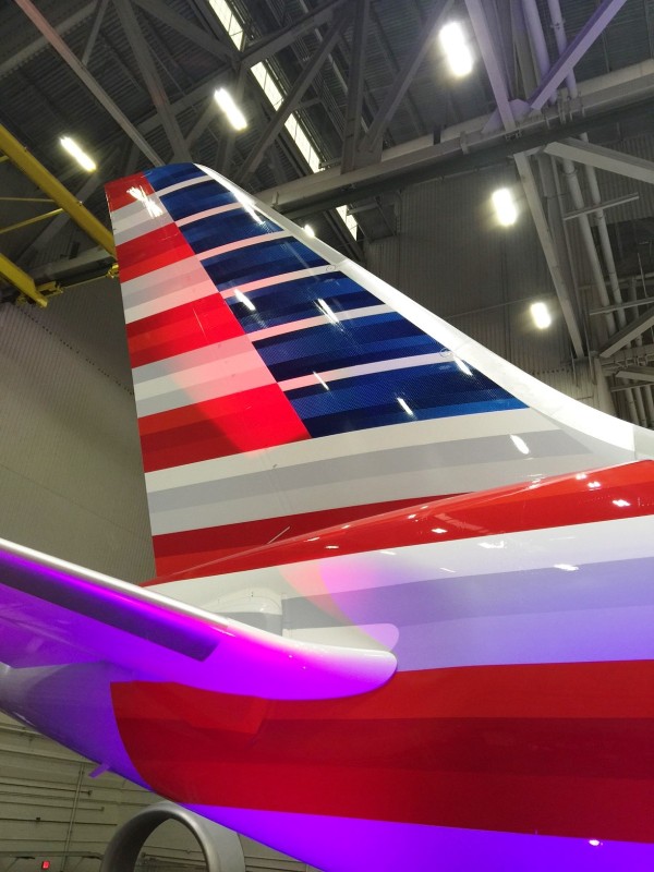 The image shows the tail section of an airplane inside a hangar. The tail is painted with horizontal stripes in red, white, and blue colors. The hangar's ceiling and lighting fixtures are visible in the background.