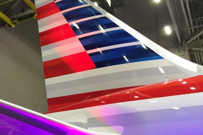 The image shows the tail section of an airplane inside a hangar. The tail is painted with horizontal stripes in red, white, and blue colors. The hangar's ceiling and lighting fixtures are visible in the background.