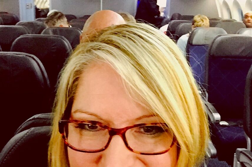 A woman with blonde hair and glasses is taking a selfie inside an airplane. She is smiling and seated in what appears to be the economy class section. Other passengers can be seen seated and standing in the background, and the overhead compartments are open with some luggage visible. The interior of the airplane is well-lit.