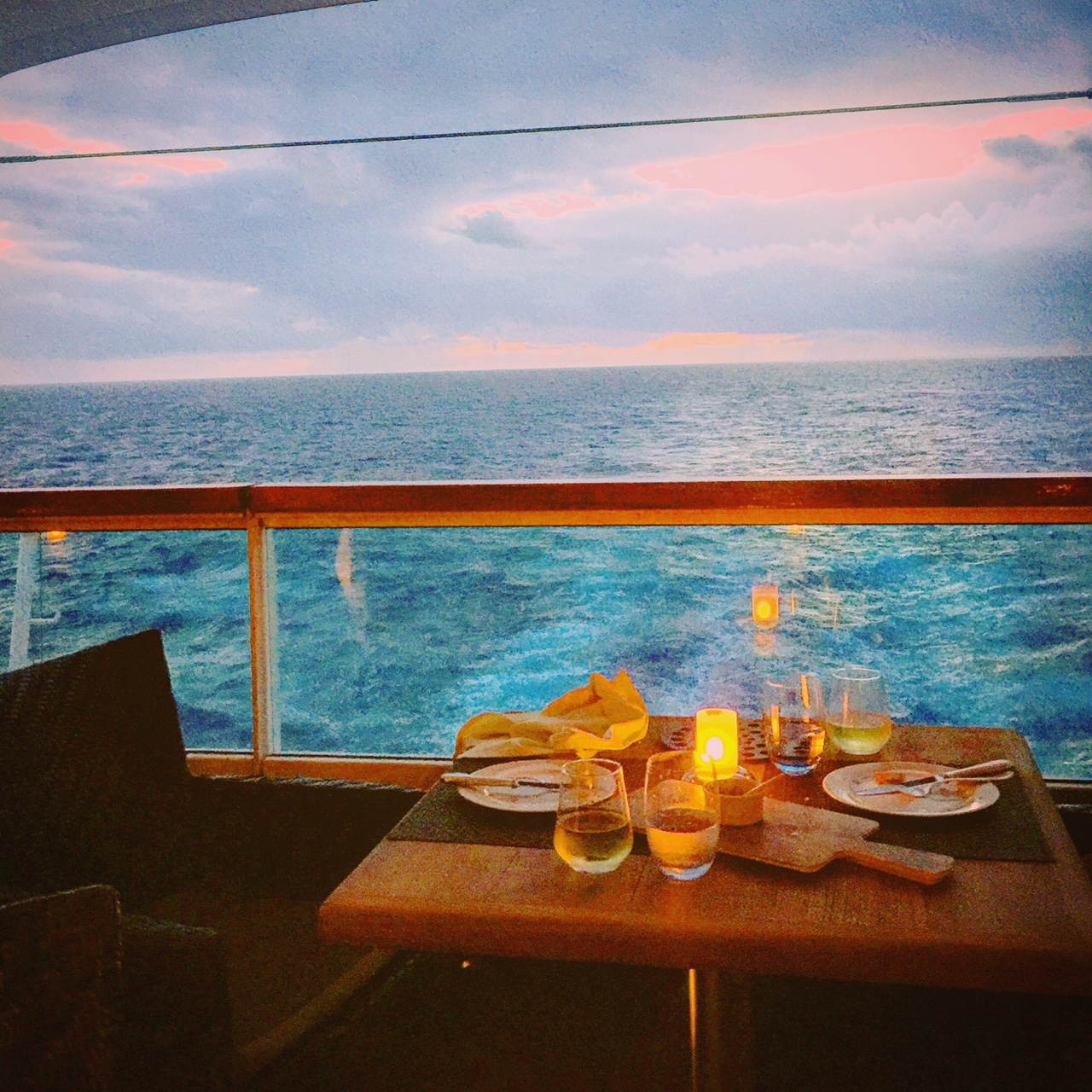 A small table set for two with plates, glasses, and a candle is positioned on a balcony overlooking the ocean. The sky is partly cloudy with hints of pink and orange from the setting or rising sun, creating a serene and romantic atmosphere. The water below is a deep blue, and the scene suggests a peaceful dining experience with a beautiful view.