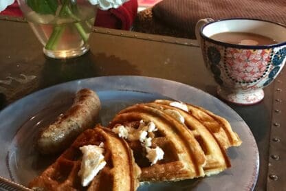A plate of breakfast food is on a table. The plate contains a sausage and several pieces of waffle with butter on top. A fork is placed on the plate. Next to the plate is a decorative cup filled with coffee or tea. In the background, there is a vase with white and pink flowers. The setting appears to be a cozy indoor environment with a couch and pillows visible.