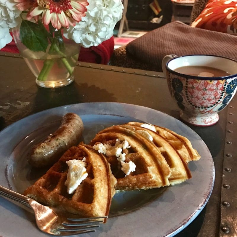 A plate of breakfast food is on a table. The plate contains a sausage and several pieces of waffle with butter on top. A fork is placed on the plate. Next to the plate is a decorative cup filled with coffee or tea. In the background, there is a vase with white and pink flowers. The setting appears to be a cozy indoor environment with a couch and pillows visible.