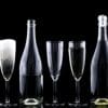 Alt text: A row of seven champagne flutes and two bottles are arranged against a black background. The first glass is being filled with a clear liquid, the second glass is filled with a foamy beverage, the third glass is empty, the fourth glass contains a small amount of liquid, the fifth glass is empty, the sixth glass is empty, and the seventh glass is empty. The two bottles, one between the third and fourth glasses and the other between the fifth and sixth glasses, are partially filled with a light-colored liquid.