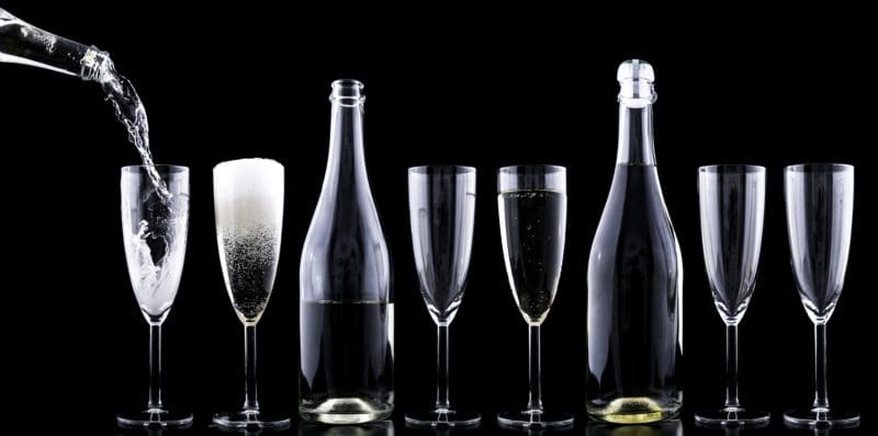 Alt text: A row of seven champagne flutes and two bottles are arranged against a black background. The first glass is being filled with a clear liquid, the second glass is filled with a foamy beverage, the third glass is empty, the fourth glass contains a small amount of liquid, the fifth glass is empty, the sixth glass is empty, and the seventh glass is empty. The two bottles, one between the third and fourth glasses and the other between the fifth and sixth glasses, are partially filled with a light-colored liquid.