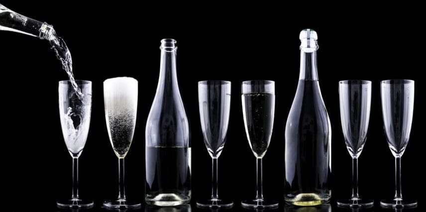 Alt text: A row of seven champagne flutes and two bottles are arranged against a black background. The first glass is being filled with a clear liquid, the second glass is filled with a foamy beverage, the third glass is empty, the fourth glass contains a small amount of liquid, the fifth glass is empty, the sixth glass is empty, and the seventh glass is empty. The two bottles, one between the third and fourth glasses and the other between the fifth and sixth glasses, are partially filled with a light-colored liquid.