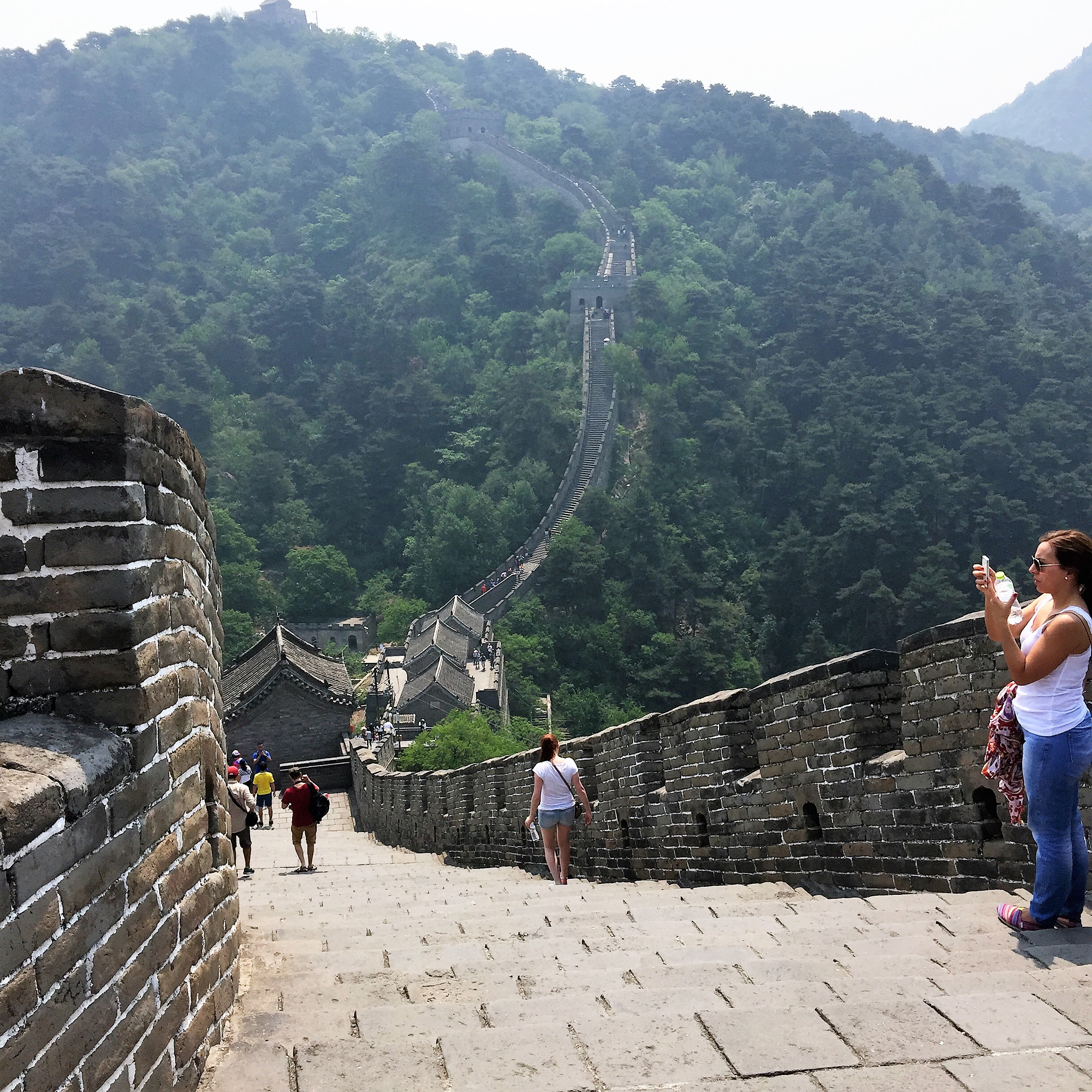 The image shows a section of the Great Wall of China, with a few tourists walking along the wall. The wall stretches into the distance, winding over the lush, green hills. One person is standing on the right side, taking a photo with a smartphone. The weather appears to be clear, providing a good view of the surrounding landscape.